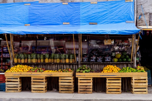 A vibrant fruit stand displays a variety of fresh fruits neatly arranged in wooden crates. The stand is covered with a blue tarp and features an array of pineapples, mangoes, oranges, and limes. In the background, packaged goods are visible, adding to the assortment of items available for sale.