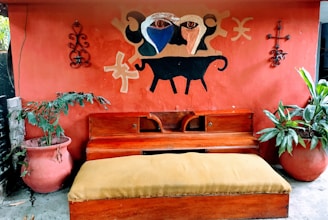 An outdoor scene features a red wall with a decorative mural depicting abstract shapes and a black animal figure. Two large terracotta pots with green plants flank a wooden bench with a mustard-colored cushion. Metal wall art pieces are on both sides of the mural.