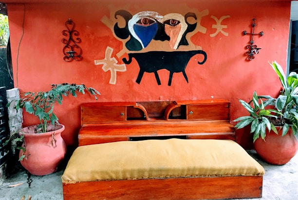An outdoor scene features a red wall with a decorative mural depicting abstract shapes and a black animal figure. Two large terracotta pots with green plants flank a wooden bench with a mustard-colored cushion. Metal wall art pieces are on both sides of the mural.