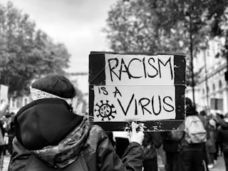 A person in a crowd holds a sign that reads 'Racism is a Virus'. The scene is monochromatic with trees and buildings in the background, suggesting an outdoor protest or demonstration.