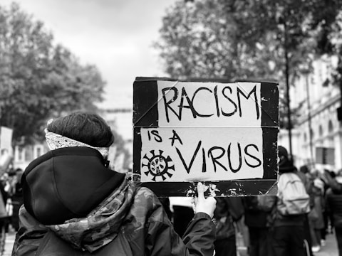 A person in a crowd holds a sign that reads 'Racism is a Virus'. The scene is monochromatic with trees and buildings in the background, suggesting an outdoor protest or demonstration.