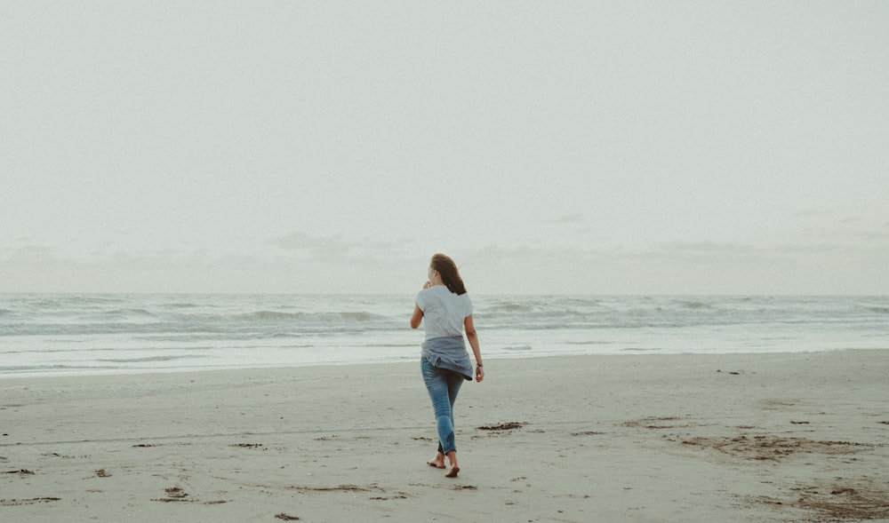 woman in white shirt and blue denim jeans walking on beach during daytime