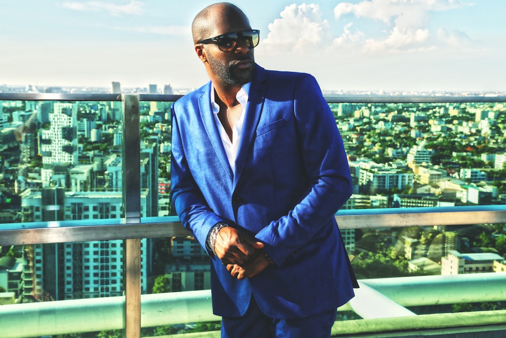 man in blue suit jacket and black sunglasses standing near green metal railings during daytime