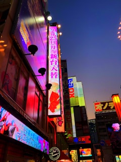A vibrant urban cityscape filled with bright, colorful neon signs against a twilight sky. The signs contain a mix of Japanese characters and bold, illuminated advertisements on tall buildings.