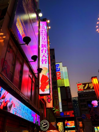 A vibrant urban cityscape filled with bright, colorful neon signs against a twilight sky. The signs contain a mix of Japanese characters and bold, illuminated advertisements on tall buildings.
