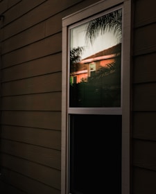 A reflection of a house with a red roof can be seen in a window on the exterior of a building. The window is surrounded by horizontal wooden siding, and there are palm tree leaves visible in the reflection.