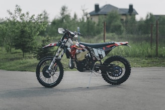 A motocross bike stands on a paved surface, surrounded by greenery. The bike, adorned with a graphic design in black, red, and white, appears rugged and well-equipped for off-road conditions. In the background, a house and trees are visible, slightly blurred to emphasize the focus on the bike.
