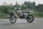 A motocross bike stands on a paved surface, surrounded by greenery. The bike, adorned with a graphic design in black, red, and white, appears rugged and well-equipped for off-road conditions. In the background, a house and trees are visible, slightly blurred to emphasize the focus on the bike.