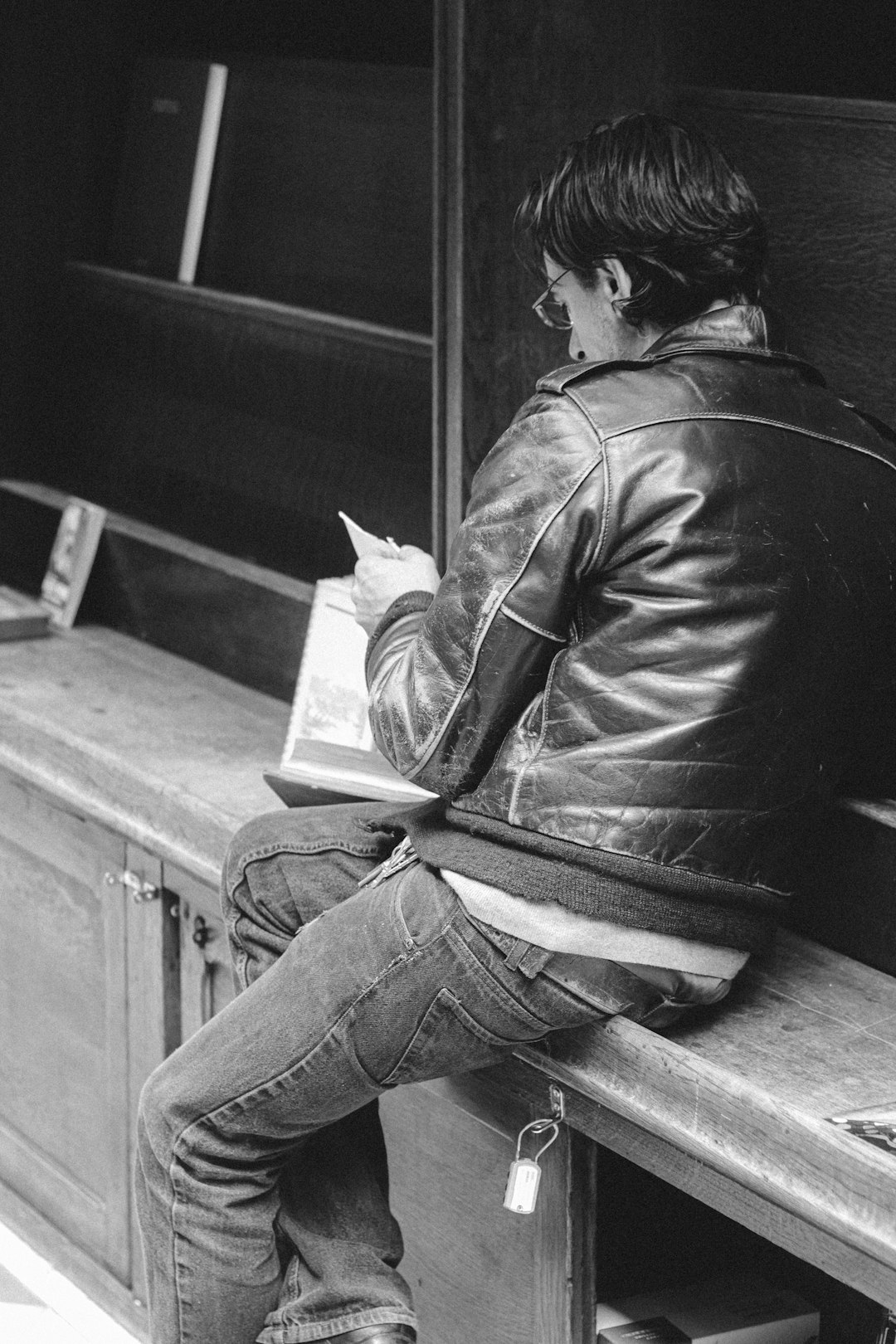 grayscale photo of woman in jacket and denim jeans sitting on wooden table reading book