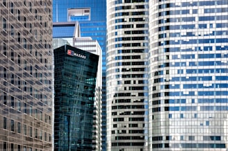 A collection of modern skyscrapers with a mix of glass and steel facades. The buildings exhibit a variety of geometric patterns, with one tower displaying a prominent logo. The reflective surfaces create a mosaic of light and shadow across the structures.