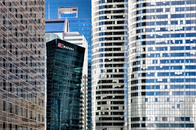 A collection of modern skyscrapers with a mix of glass and steel facades. The buildings exhibit a variety of geometric patterns, with one tower displaying a prominent logo. The reflective surfaces create a mosaic of light and shadow across the structures.