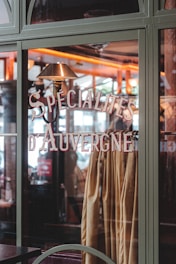 A glass window showcases the words 'Specialites d'Auvergne' in stylish lettering. Inside, a warm, cozy interior is visible with a soft, ambient light that highlights wood elements and warm colors. Curtains or fabric in earthy tones hang behind the window glass.