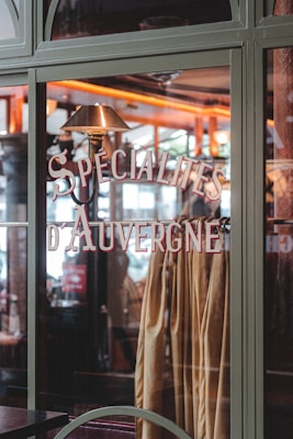 A glass window showcases the words 'Specialites d'Auvergne' in stylish lettering. Inside, a warm, cozy interior is visible with a soft, ambient light that highlights wood elements and warm colors. Curtains or fabric in earthy tones hang behind the window glass.