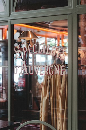A glass window showcases the words 'Specialites d'Auvergne' in stylish lettering. Inside, a warm, cozy interior is visible with a soft, ambient light that highlights wood elements and warm colors. Curtains or fabric in earthy tones hang behind the window glass.
