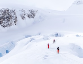 Three mountaineers with backpacks and ski poles traverse a snowy, mountainous landscape. The scene includes snow-covered peaks and crevices, with prominent jagged rocks visible in the background.