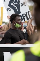 A group of people participate in a protest, with signs bearing messages. Many are wearing face masks, and one prominent sign features a raised fist symbol and words advocating change.