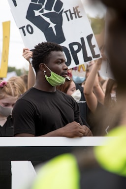 A group of people participate in a protest, with signs bearing messages. Many are wearing face masks, and one prominent sign features a raised fist symbol and words advocating change.