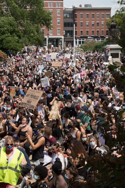 A large crowd of people gathered in an urban area, holding signs and wearing face masks. The crowd appears diverse and is situated in front of brick buildings and greenery. A statue is visible to the right, and many participants are engaged and focused, creating a sense of solidarity.