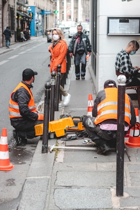 Two workers wearing reflective vests are kneeling on the sidewalk, engaged in maintenance work. They are surrounded by orange traffic cones and various equipment. A woman in a bright orange coat and white mask walks by, and another pedestrian in a black jacket approaches from the background.