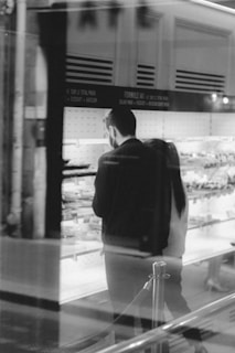 A black and white photograph of two people standing in front of a display case with various items. The setting appears to be a store or a bakery with shelves and signage visible in the background. The people are seen through a glass window, adding a reflective quality to the image.
