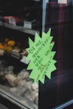A green star-shaped note with Korean text is attached to a glass door of a refrigerator in a store. Inside the refrigerator, there are various fresh produce items, including vegetables and possibly fruits, stored on metal racks.