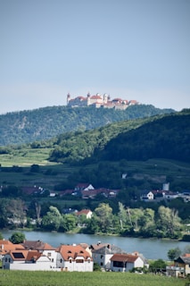 A picturesque landscape featuring a hilltop monastery with red rooftops and towers, nestled among lush green forests. Below the hill lies a tranquil village with houses that have red and gray roofs. A calm river runs in front of the village, reflecting the serene atmosphere.