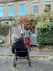 A person in a wheelchair is holding up a smartphone, possibly taking a photo or video of another person standing nearby. The standing person is dressed casually in red pants and a gray jacket. Behind them is a house with a stone facade and several large windows, surrounded by vibrant greenery and blooming red flowers.