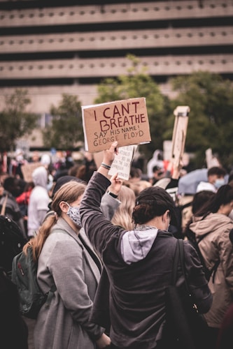 A group of people gathered in a protest, one individual prominently holding a sign that reads 'I CAN'T BREATHE, SAY HIS NAME! GEORGE FLOYD'. The crowd appears diverse, with many wearing face masks. The background includes trees and a large building, suggesting an urban setting.