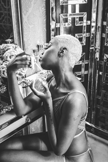 A person with short hair and wearing lingerie sits at a vanity, holding a perfume bottle with one hand and a dropper in the other. The vanity is adorned with decorative elements, including flowers and reflective surfaces, creating an elegant and intimate setting.