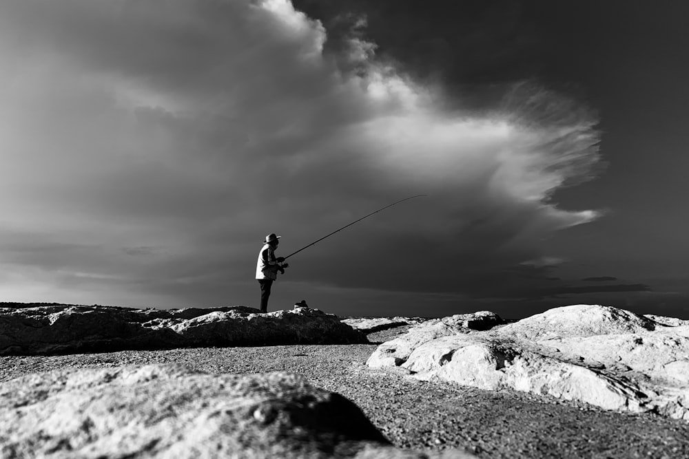 Foto en escala de grises de un hombre con chaqueta negra y pantalones sosteniendo una caña de pescar