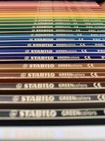 A close-up perspective of a series of colored pencils aligned in parallel. Each pencil is labeled with 'STABILO GREENcolors' and the image features a gradient of colors ranging from earthy tones to vibrant hues.