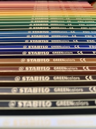 A close-up perspective of a series of colored pencils aligned in parallel. Each pencil is labeled with 'STABILO GREENcolors' and the image features a gradient of colors ranging from earthy tones to vibrant hues.