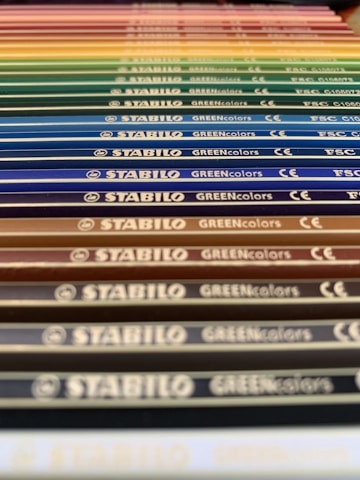 A close-up perspective of a series of colored pencils aligned in parallel. Each pencil is labeled with 'STABILO GREENcolors' and the image features a gradient of colors ranging from earthy tones to vibrant hues.