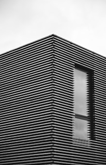 A modern building facade with a window, featuring horizontal slats creating a striped pattern. The overall appearance is minimalist and sleek.