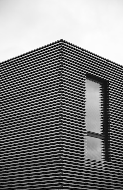 A modern building facade with a window, featuring horizontal slats creating a striped pattern. The overall appearance is minimalist and sleek.