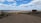 A vast, arid landscape with a dirt road leading towards a distant horizon. Sparse vegetation is scattered throughout the area. A sign on the right side reads 'Bajo de la Laguna Grande Bosques Petrificados'. The sky is expansive, filled with scattered clouds and patches of blue.