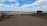 A vast, arid landscape with a dirt road leading towards a distant horizon. Sparse vegetation is scattered throughout the area. A sign on the right side reads 'Bajo de la Laguna Grande Bosques Petrificados'. The sky is expansive, filled with scattered clouds and patches of blue.