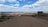A vast, arid landscape with a dirt road leading towards a distant horizon. Sparse vegetation is scattered throughout the area. A sign on the right side reads 'Bajo de la Laguna Grande Bosques Petrificados'. The sky is expansive, filled with scattered clouds and patches of blue.