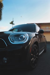 A close-up view of a sleek black car is shown from a low angle, emphasizing its glossy surface and modern headlight design. The lighting suggests a late afternoon or early evening setting, with a clear blue sky serving as a backdrop. The image captures the front and side of the car, highlighting its shiny finish and detailing.