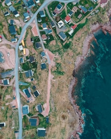 An aerial view of a coastal area with scattered houses, roads, and natural landscape. The image features a rocky shoreline with clear blue waters on one side and a small settlement of houses with roads curving through green and brown patches of land on the other.