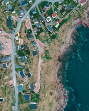 An aerial view of a coastal area with scattered houses, roads, and natural landscape. The image features a rocky shoreline with clear blue waters on one side and a small settlement of houses with roads curving through green and brown patches of land on the other.