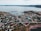 A coastal town with a collection of small houses and buildings is nestled by the seaside. There is a harbor with various docks extending into the water, and several boats are moored. The landscape is hilly, and a mixture of grassy and rocky terrain leads to the water's edge. Roads curve through the town, connecting the different areas. The scene conveys a sense of quiet and isolation, typical of a rural seaside community.