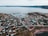 A coastal town with a collection of small houses and buildings is nestled by the seaside. There is a harbor with various docks extending into the water, and several boats are moored. The landscape is hilly, and a mixture of grassy and rocky terrain leads to the water's edge. Roads curve through the town, connecting the different areas. The scene conveys a sense of quiet and isolation, typical of a rural seaside community.
