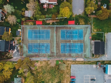 An aerial view of two adjacent outdoor tennis courts with a blue surface, surrounded by residential houses and green spaces. The scene includes nearby trees, a parking area with several cars, and pathways leading to the courts.