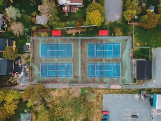 An aerial view of two adjacent outdoor tennis courts with a blue surface, surrounded by residential houses and green spaces. The scene includes nearby trees, a parking area with several cars, and pathways leading to the courts.