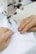 A close-up of hands guiding white fabric through a sewing machine while attaching a label. The label reads 'CHAPTER ONE DOROE'. The person is wearing rings and has blue painted nails.