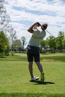 A golfer is captured mid-swing on a lush green golf course under a partly cloudy sky. The individual wears a white shirt, dark shorts, a cap, and golf shoes. Surrounding the player are trees with green foliage, and the sun casts shadows on the ground.
