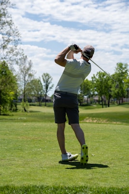 A golfer is captured mid-swing on a lush green golf course under a partly cloudy sky. The individual wears a white shirt, dark shorts, a cap, and golf shoes. Surrounding the player are trees with green foliage, and the sun casts shadows on the ground.