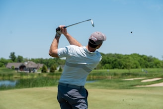 A person is playing golf on a well-manicured golf course. The individual is wearing a cap, gloves, and casual golfing attire while holding a golf club in a follow-through swing posture. In the background, there are grassy areas, a body of water, and a distant building, set under a clear blue sky.