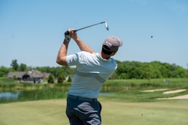 A person is playing golf on a well-manicured golf course. The individual is wearing a cap, gloves, and casual golfing attire while holding a golf club in a follow-through swing posture. In the background, there are grassy areas, a body of water, and a distant building, set under a clear blue sky.